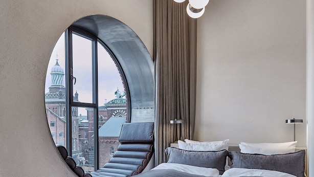 A bedroom with a large circular window, a bed, a chair, and a view of a cityscape. - Boutique Hotel Ottilia (Copenhagen)