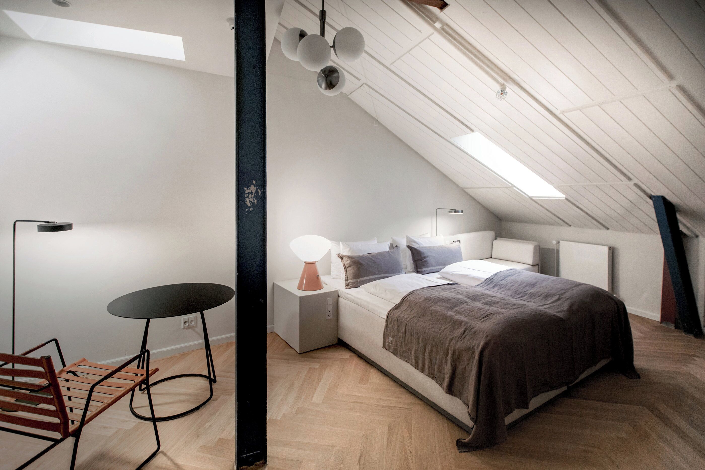 Un dormitorio moderno con techo inclinado, una cama con manta marrón, una mesita redonda con lámpara y una silla de madera.