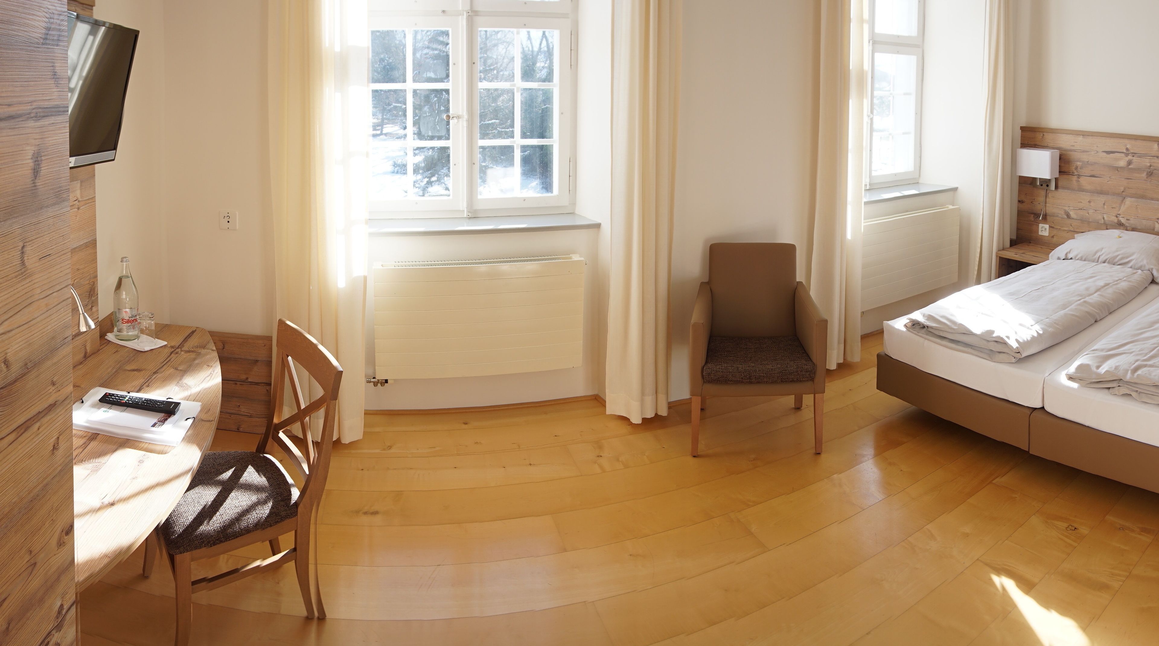 A room with a bed, a chair, a desk with a remote control, a television, and a window with curtains.