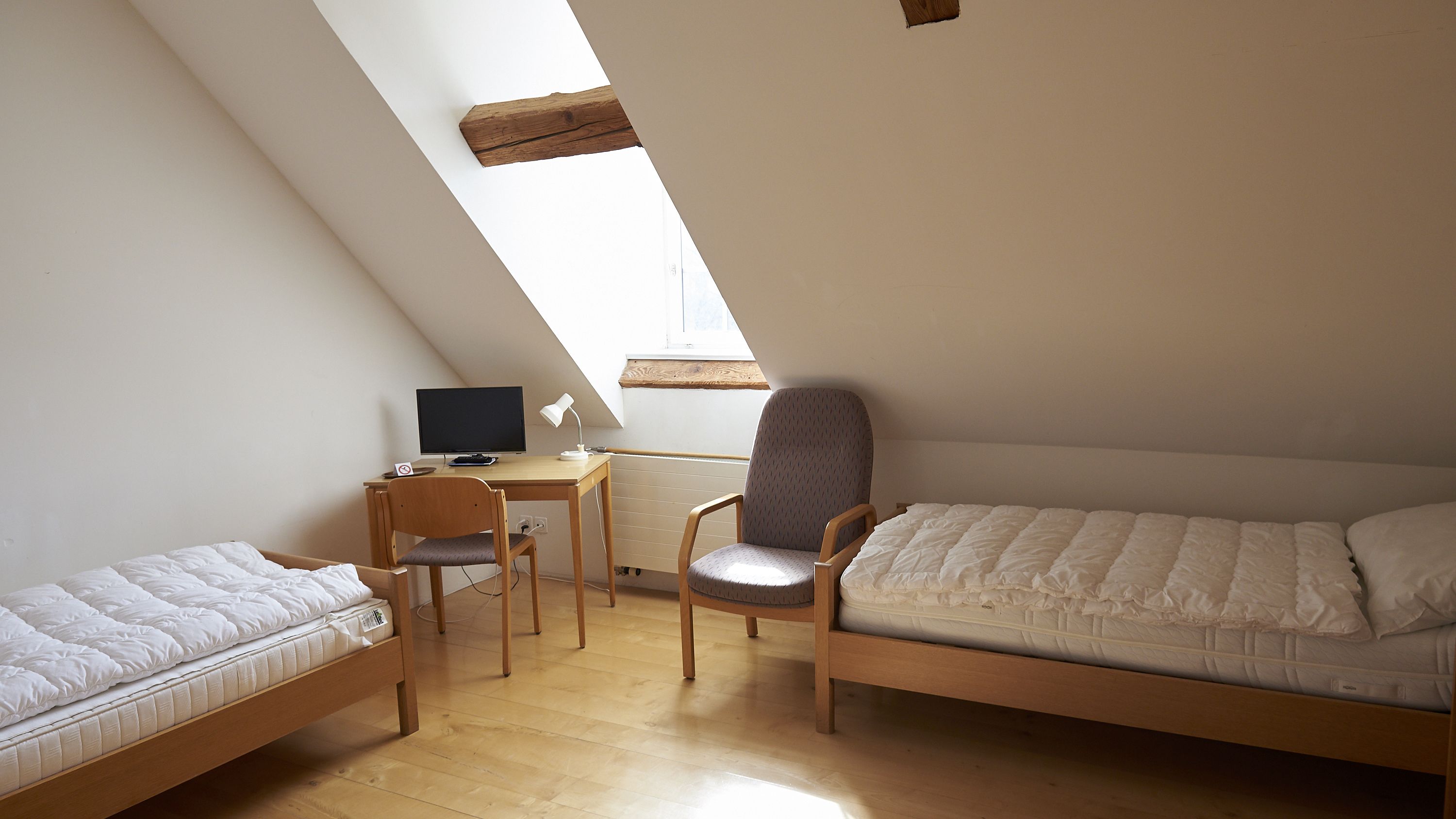 Una habitación pequeña y bien iluminada, con techo inclinado, un escritorio con computadora y silla, y dos camas individuales.
