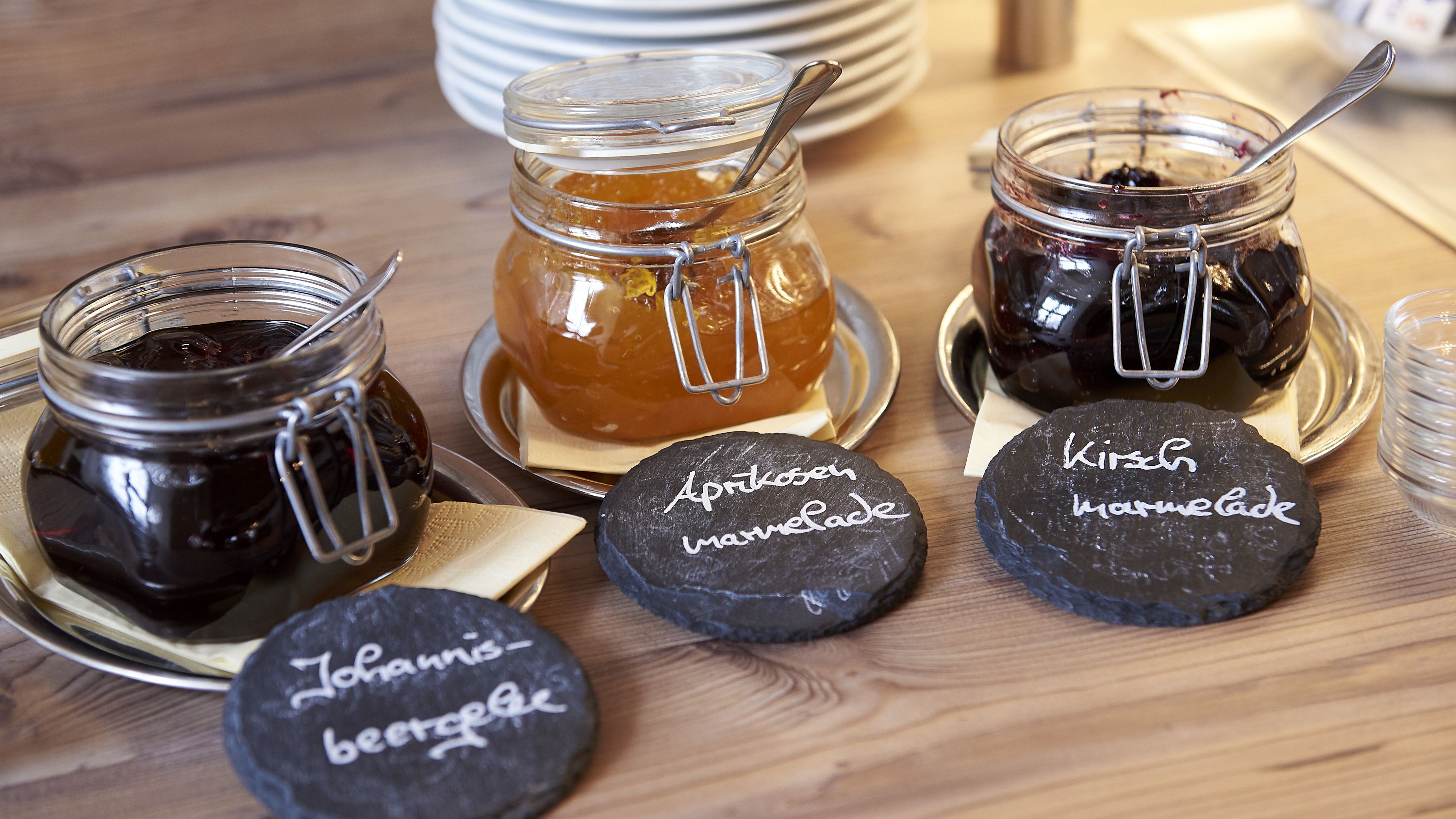 Three jars of preserves and two cheese wheels on a wooden table.