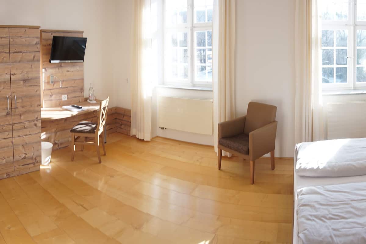 A bedroom with a bed, a desk with a chair, a TV mounted on a wooden cabinet, and a window with curtains.