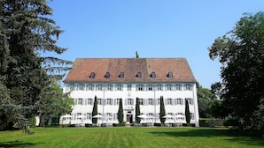 A large, white building with a red-tiled roof, surrounded by a well-maintained lawn and tall trees. - Klosterhotel Kreuz (Metzerlen)