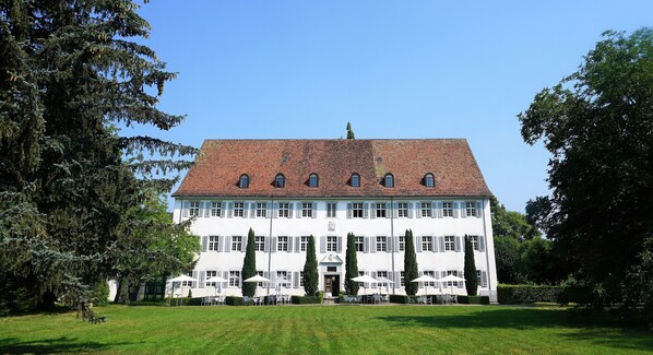 A large, white building with a red-tiled roof, surrounded by a well-maintained lawn and tall trees. - Klosterhotel Kreuz (Metzerlen)