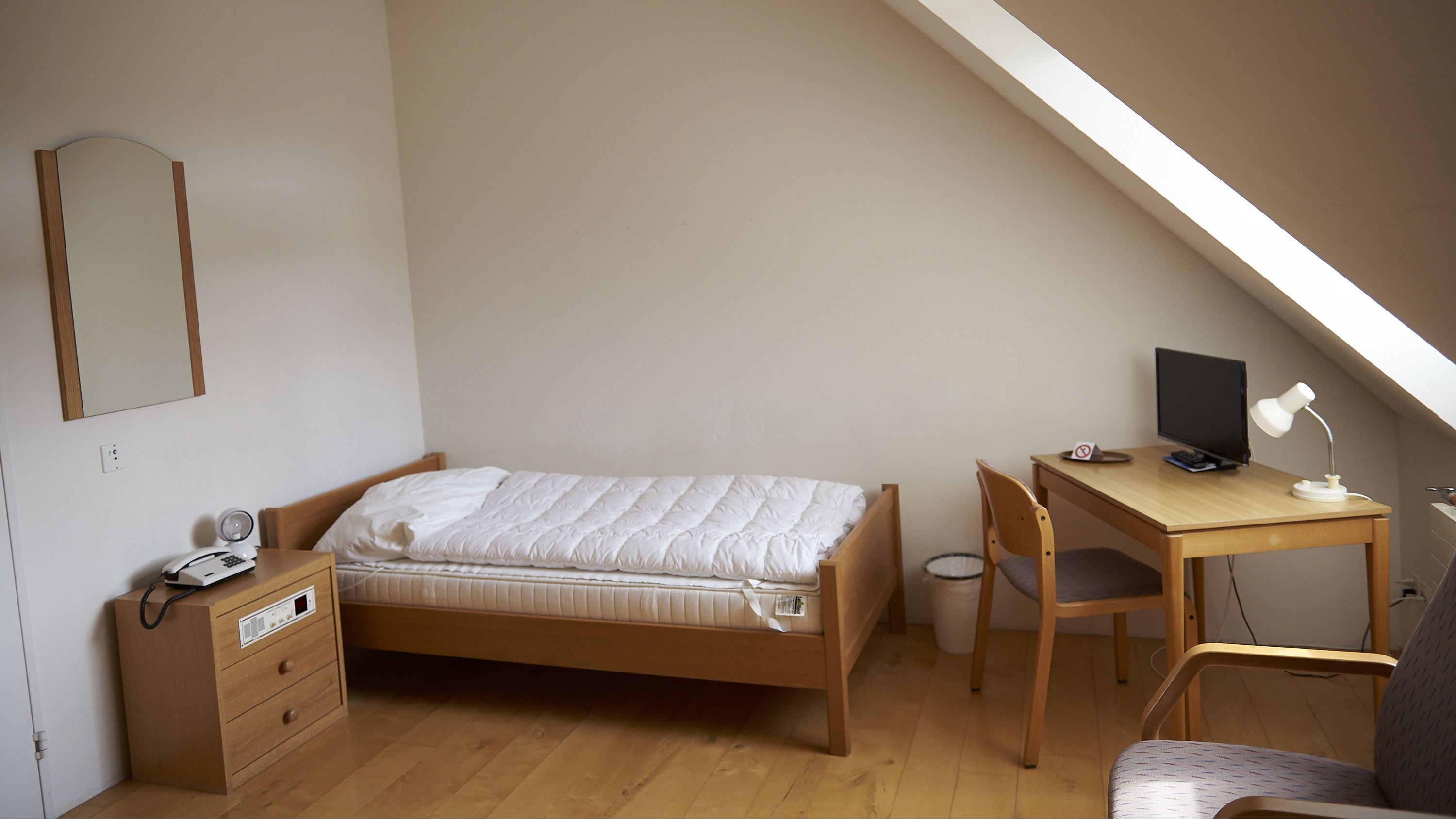 A small, tidy room with a bed, a desk, a chair, a mirror, and a small trash bin.