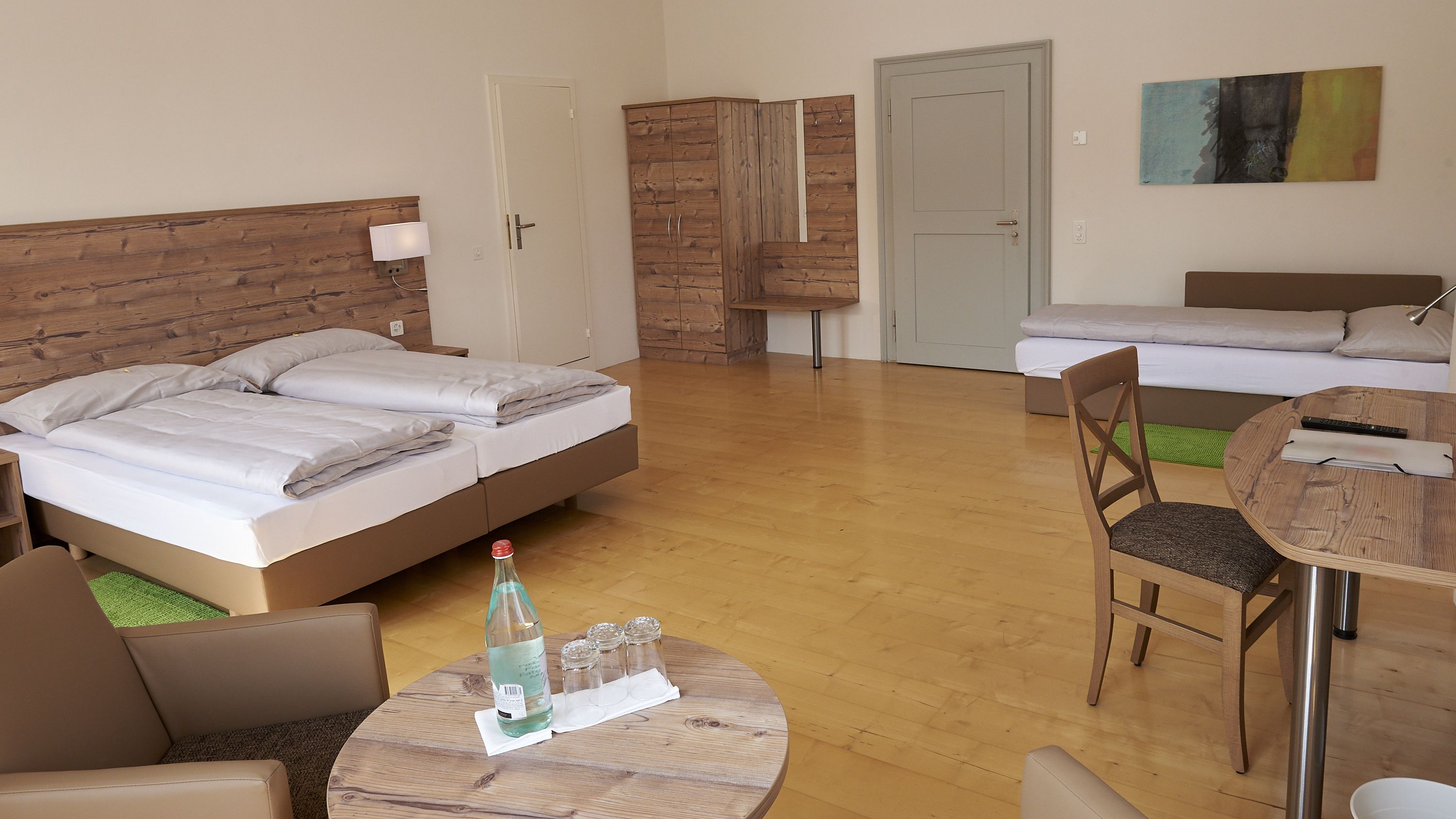 Camera d'albergo con testiera in legno, un letto con lenzuola bianche, un secondo letto, un tavolo in legno con una sedia e un quadro appeso al muro.