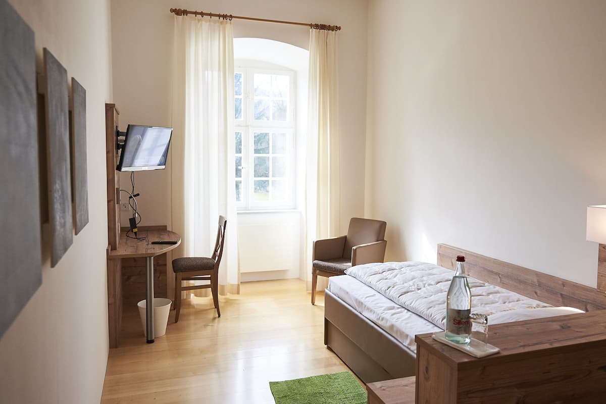 A hotel room with a bed, a desk with a TV, a chair, and a bedside table with a lamp.
