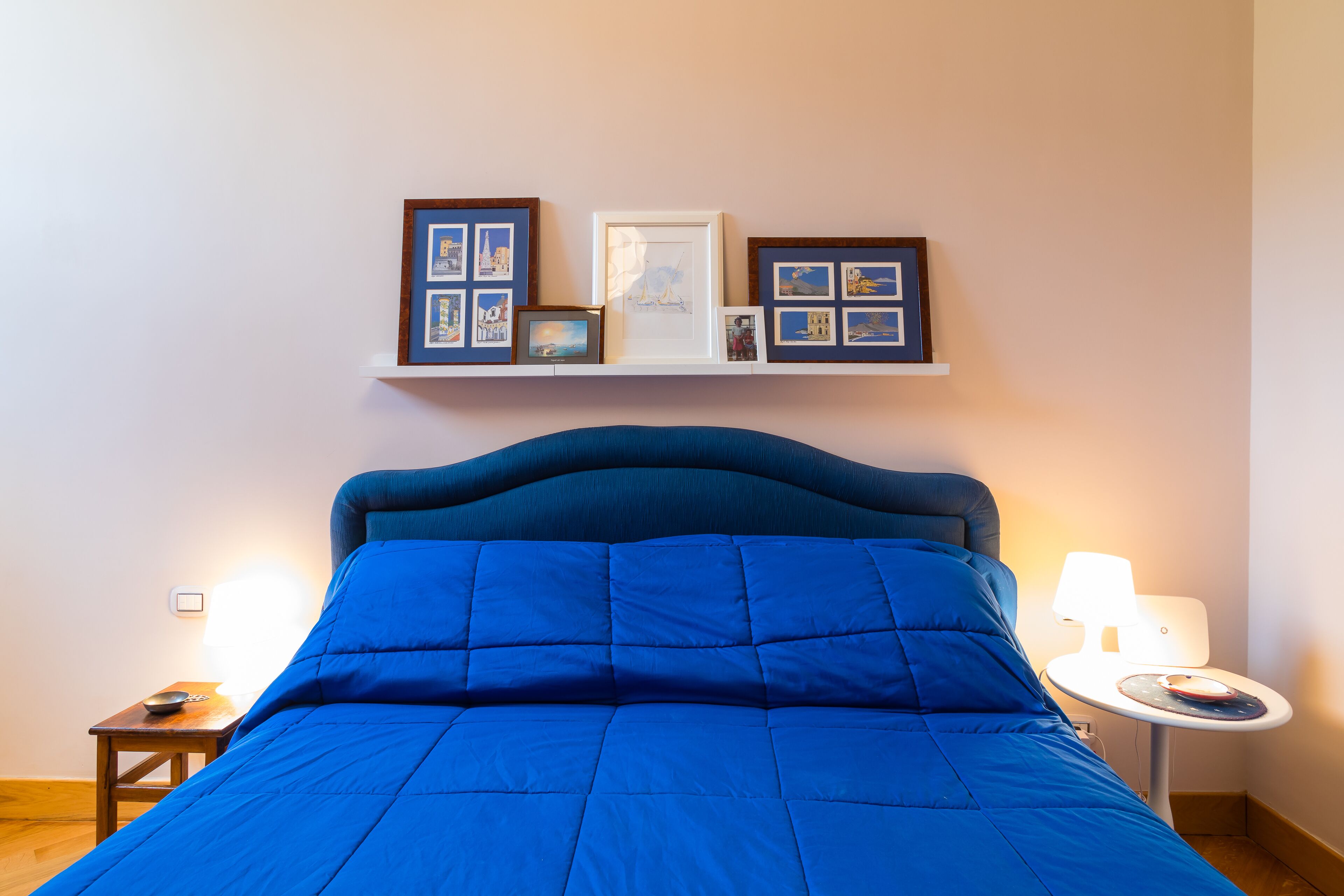 A bedroom with a large bed, a bedside table with a lamp, and a wall-mounted shelf with framed pictures.
