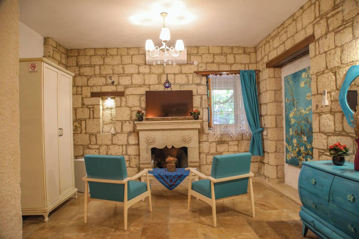 A room with stone walls, a fireplace, two teal chairs, a white cabinet, and a blue dresser.