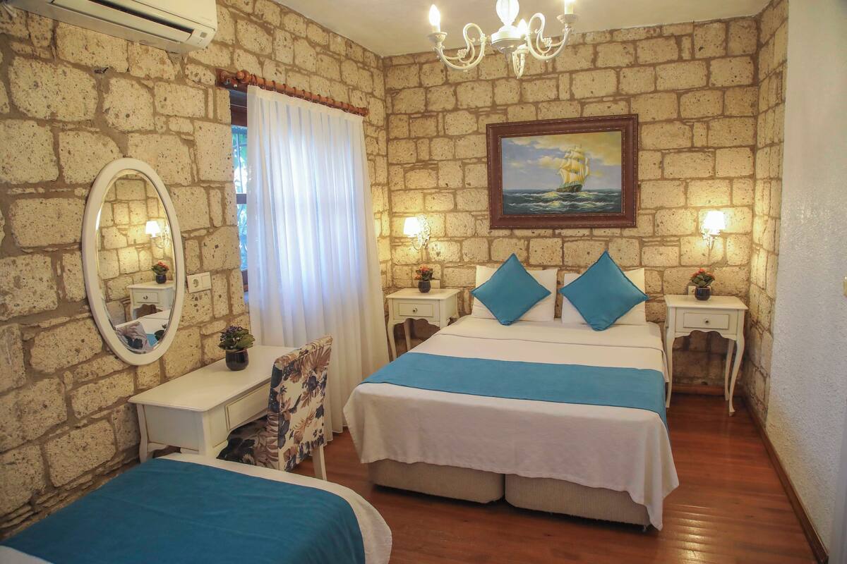 A bedroom with a brick wall, a bed with blue bedding, a bedside table with a lamp, and a painting of a ship on the wall.