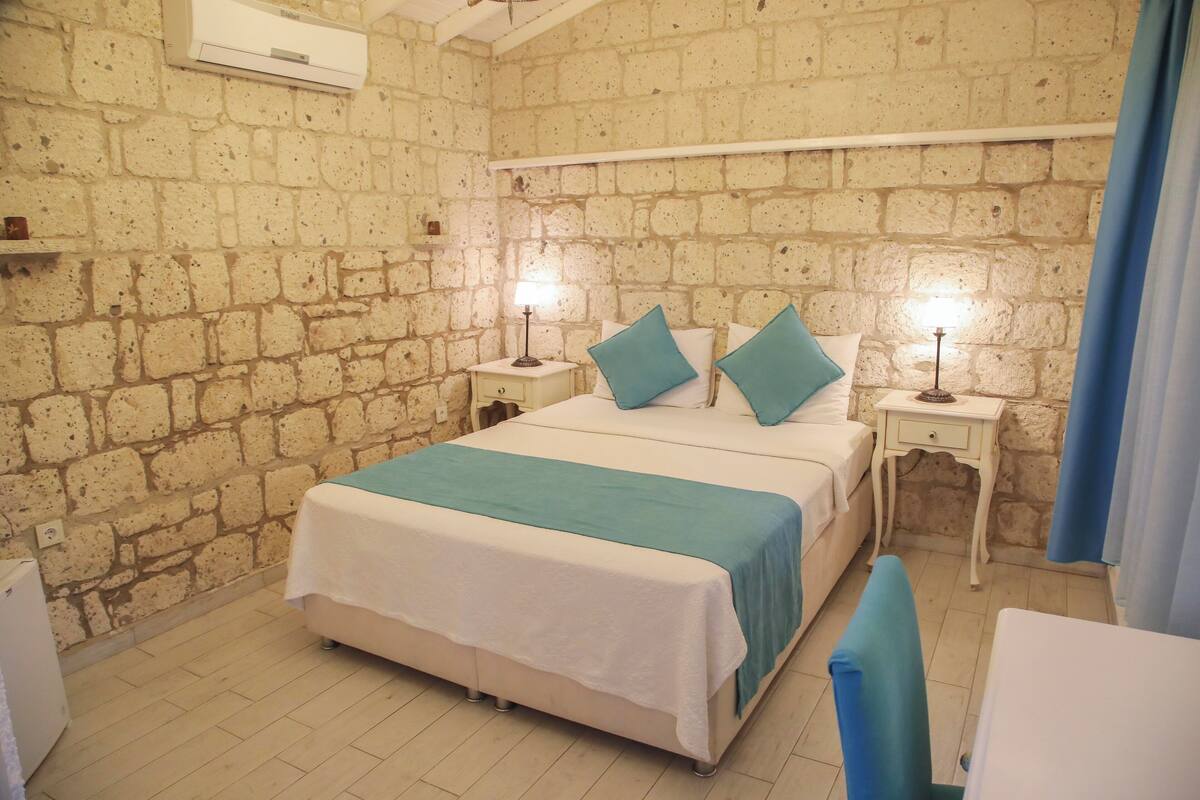 A bedroom with a brick wall, a bed with teal bedding, two bedside tables with lamps, and a small desk with a chair.