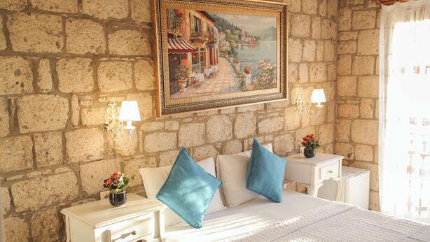 A bedroom with a stone wall, a bed with white bedding, two nightstands with lamps, and a painting of a coastal scene above the bed. - Kavalalı Butik Otel (Cesme)