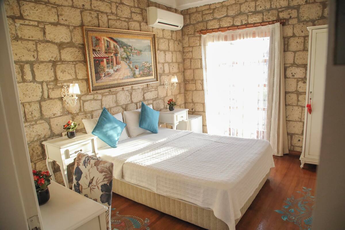 A bedroom with a stone wall, a bed with white bedding, two nightstands with lamps, a painting, and a window with curtains.