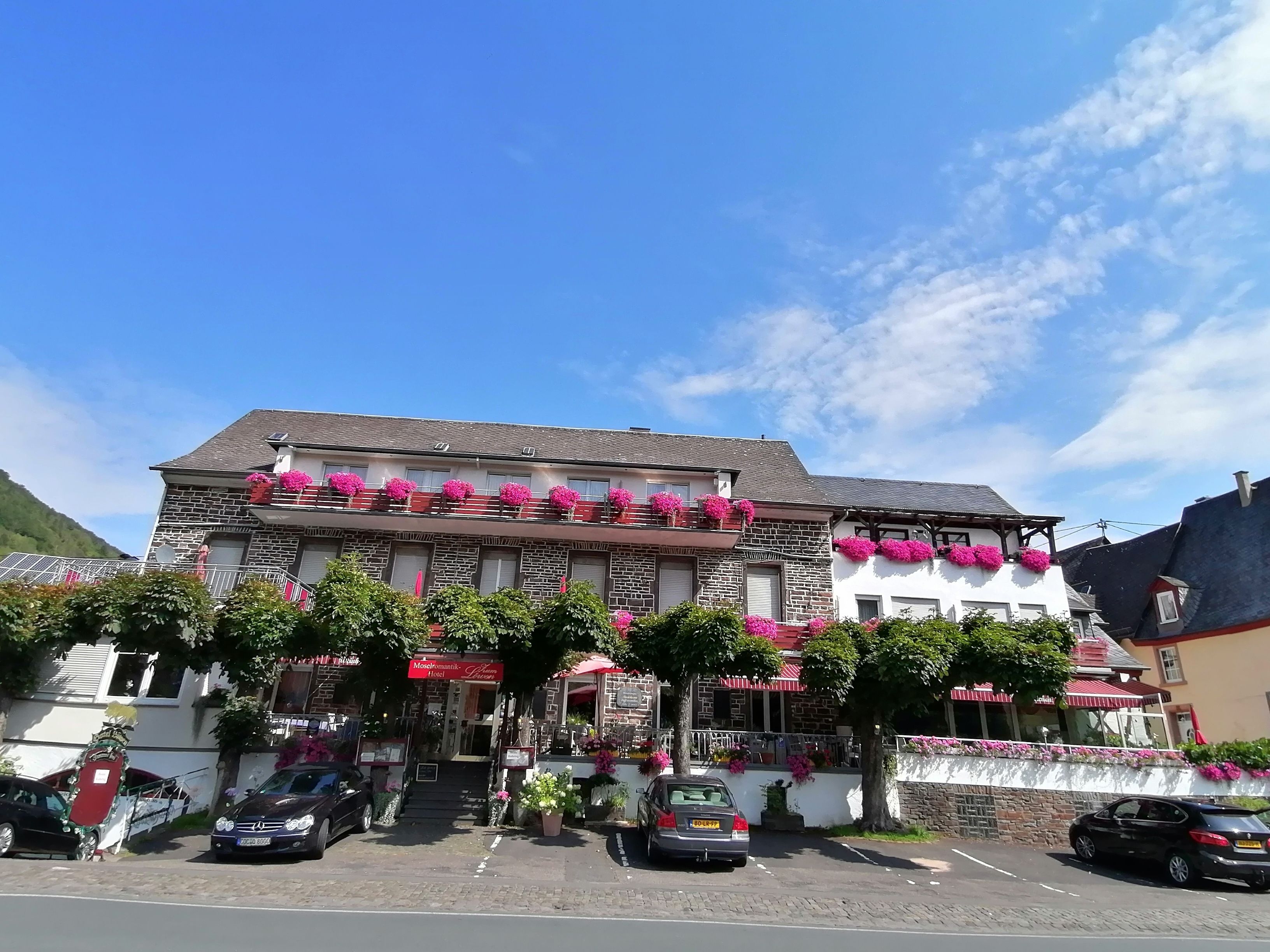 A hotel with a stone facade, multiple balconies with pink flowers, and a parking lot with cars.