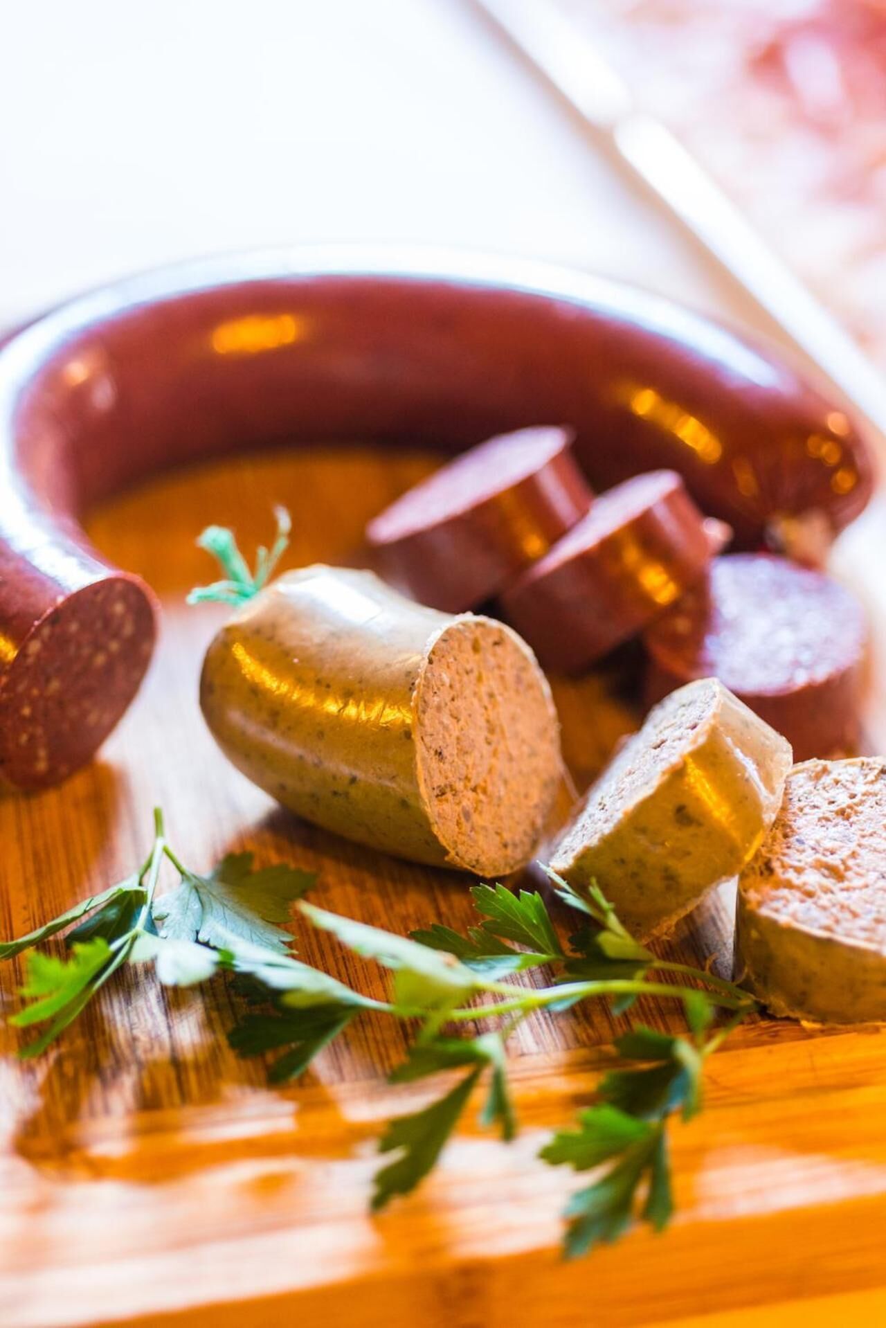 Sliced sausage and a whole sausage on a wooden surface with parsley leaves.