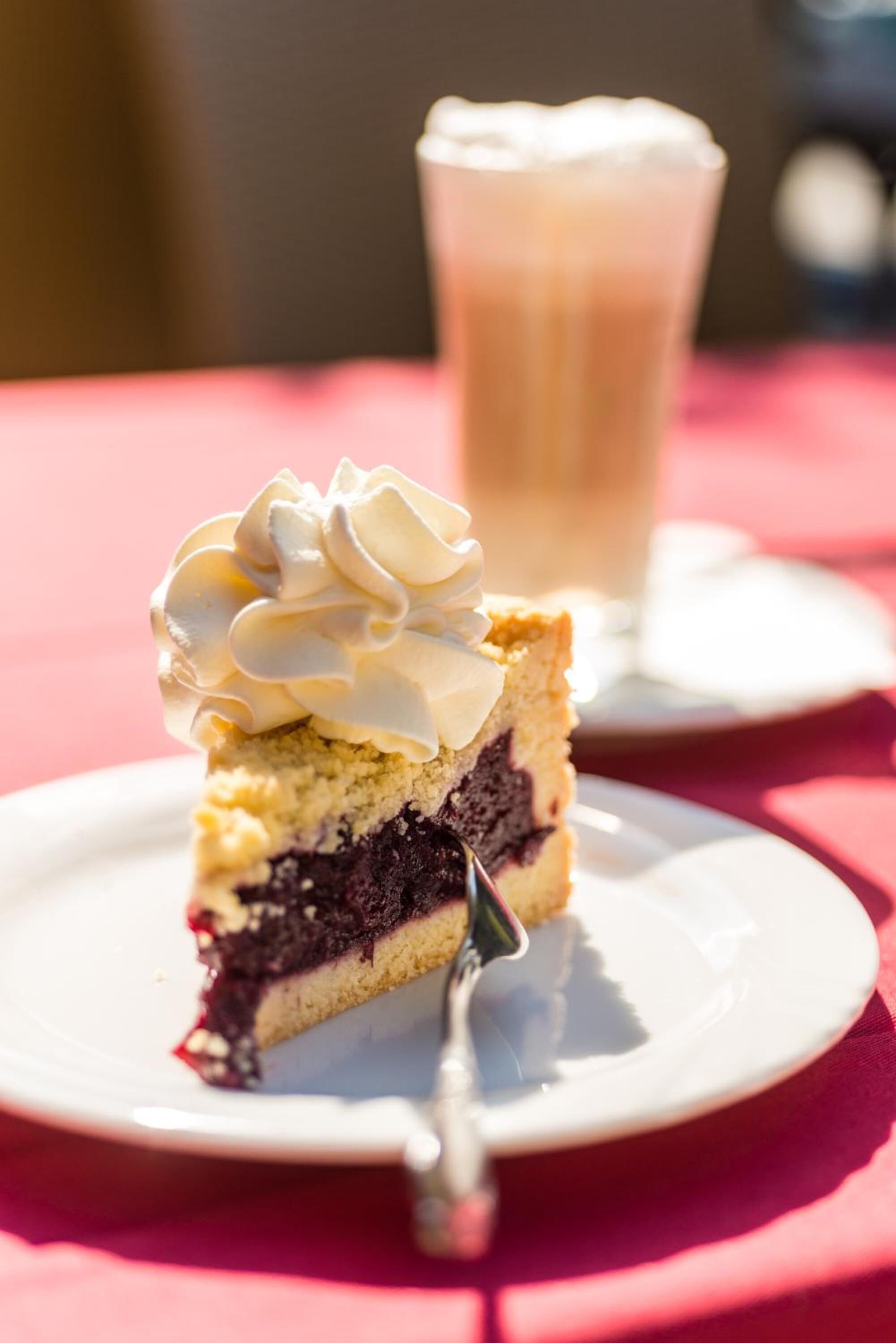 Ein Stück Kuchen mit Schlagsahne auf einem weißen Teller, im Hintergrund ein Glas helles Getränk.