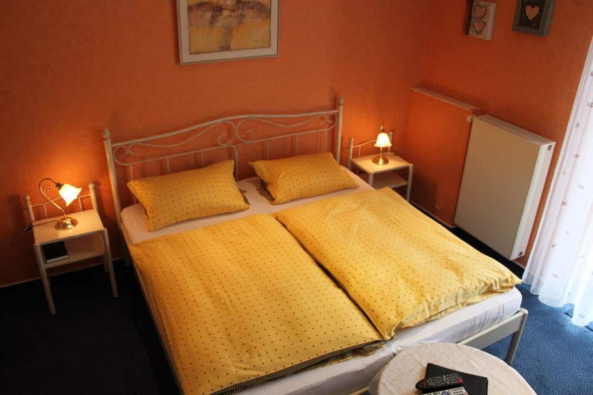 Un lit simple avec une literie jaune, deux tables de chevet avec des lampes, un radiateur et un tableau accroché au mur.