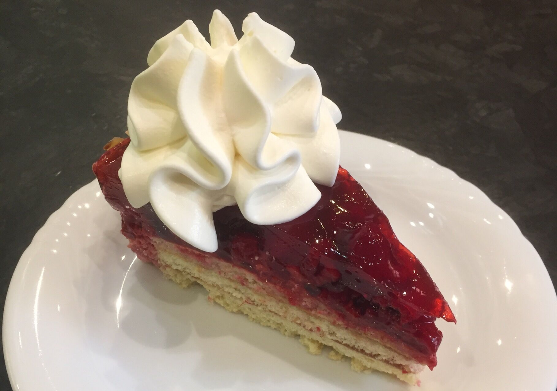 A slice of layered cake with a glossy red topping and a dollop of whipped cream on a white plate.