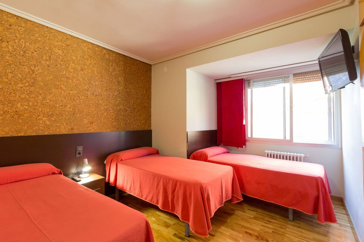 A hotel room with three beds, a television mounted on the wall, and a window with red curtains.