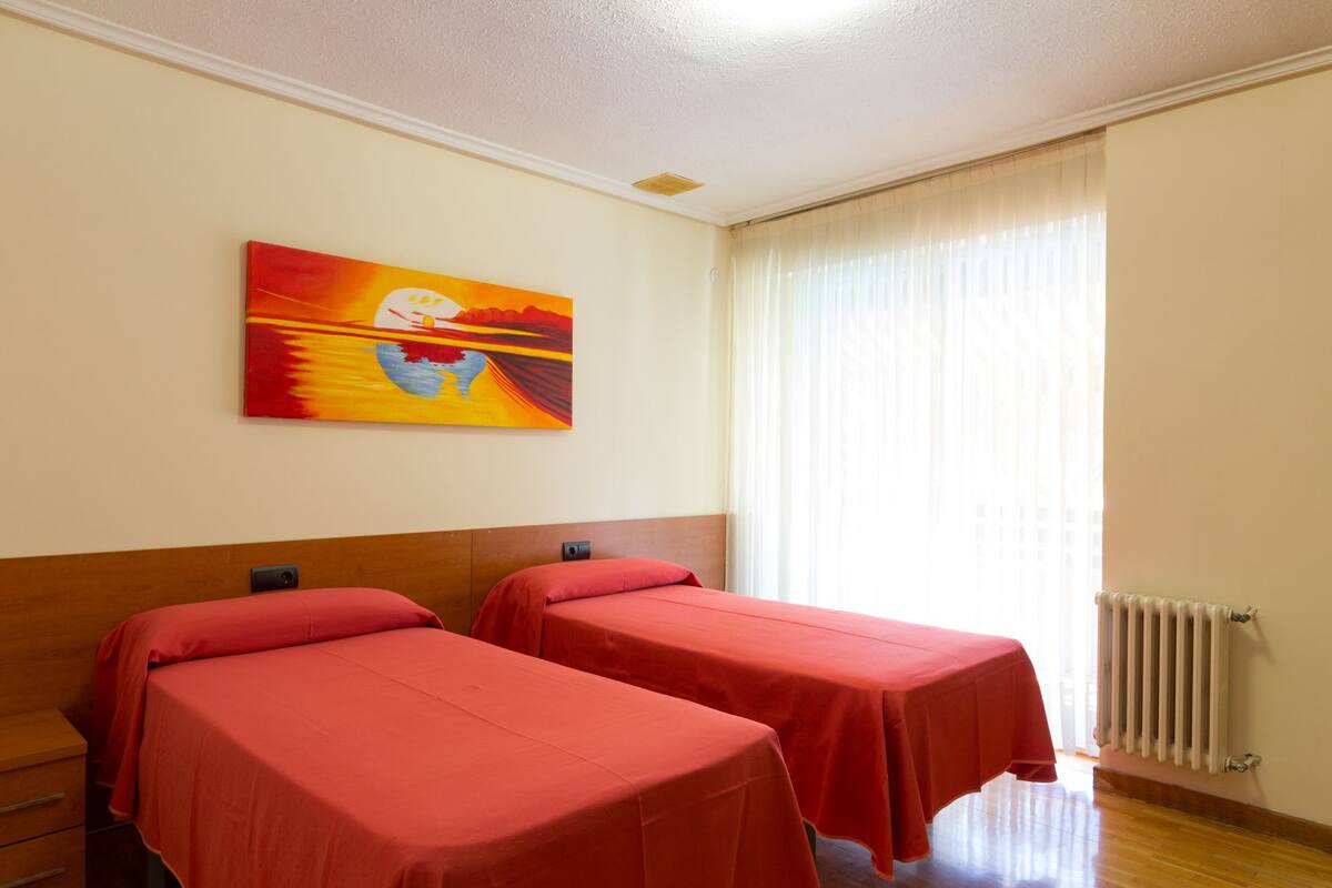A hotel room with two beds, a painting on the wall, a window with curtains, and a radiator.