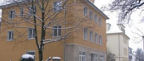 A multi-story building with a yellow facade, a tree in front, and snow on the ground.