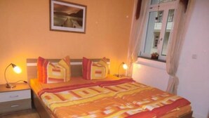 Ein Schlafzimmer mit einem Bett, zwei Nachttischlampen, einem Fenster mit Vorhängen und einem gerahmten Bild an der Wand.