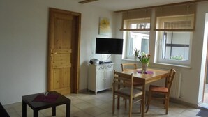 A dining area with a wooden table and chairs, a television on a cabinet, and a window with blinds. - Ferienwohnung am Kamp (Bad Doberan)