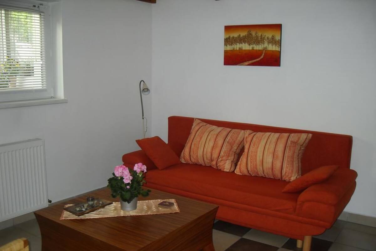A living room with a red sofa, a wooden coffee table with a vase of flowers, a painting on the wall, and a window with blinds.