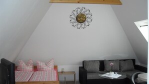 A cozy loft bedroom with a sloped ceiling, a wooden ceiling beam, a bed with pink and white bedding, a black sofa, a small table with a vase, and a wall-mounted decorative piece. - Ferienwohnung am Kamp (Bad Doberan)