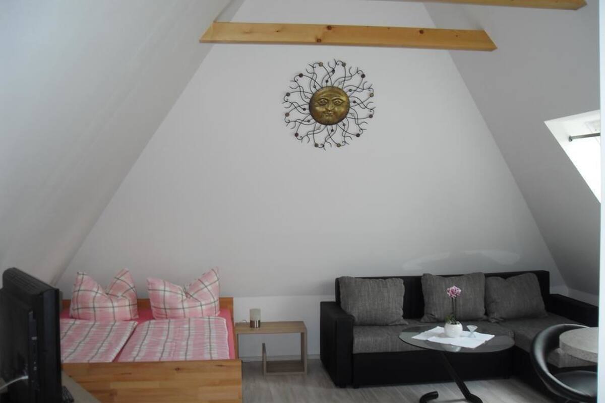 A cozy loft bedroom with a sloped ceiling, a wooden ceiling beam, a bed with pink and white bedding, a black sofa, a small table with a vase, and a wall-mounted decorative piece.