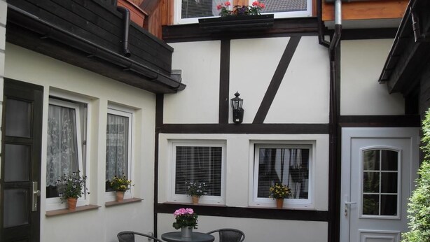 A traditional half-timbered building with a white facade and dark timber framing. There are two windows with shutters, a door, and a small outdoor seating area with a table and chairs. - Ferienwohnung am Kamp (Bad Doberan)