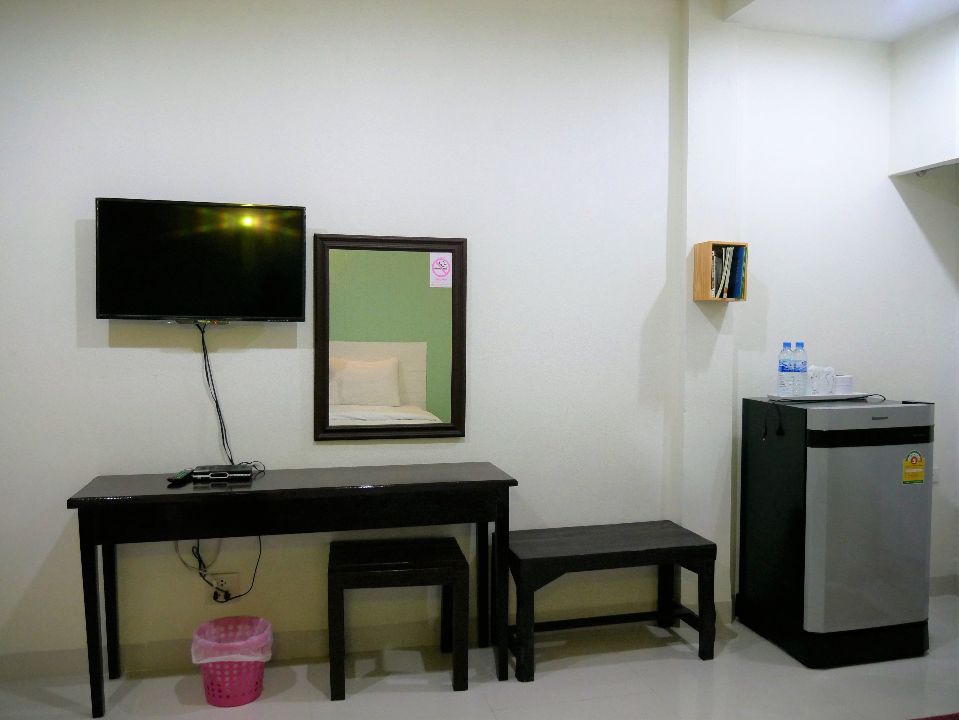 A small room with a desk, a television, a mirror, a bench, a trash bin, and a refrigerator.