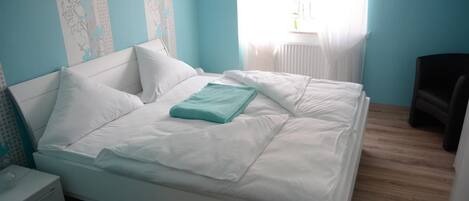 Un lit bien fait, avec une literie blanche et un plaid turquoise, dans une pièce aux murs bleu clair et au plancher en bois.