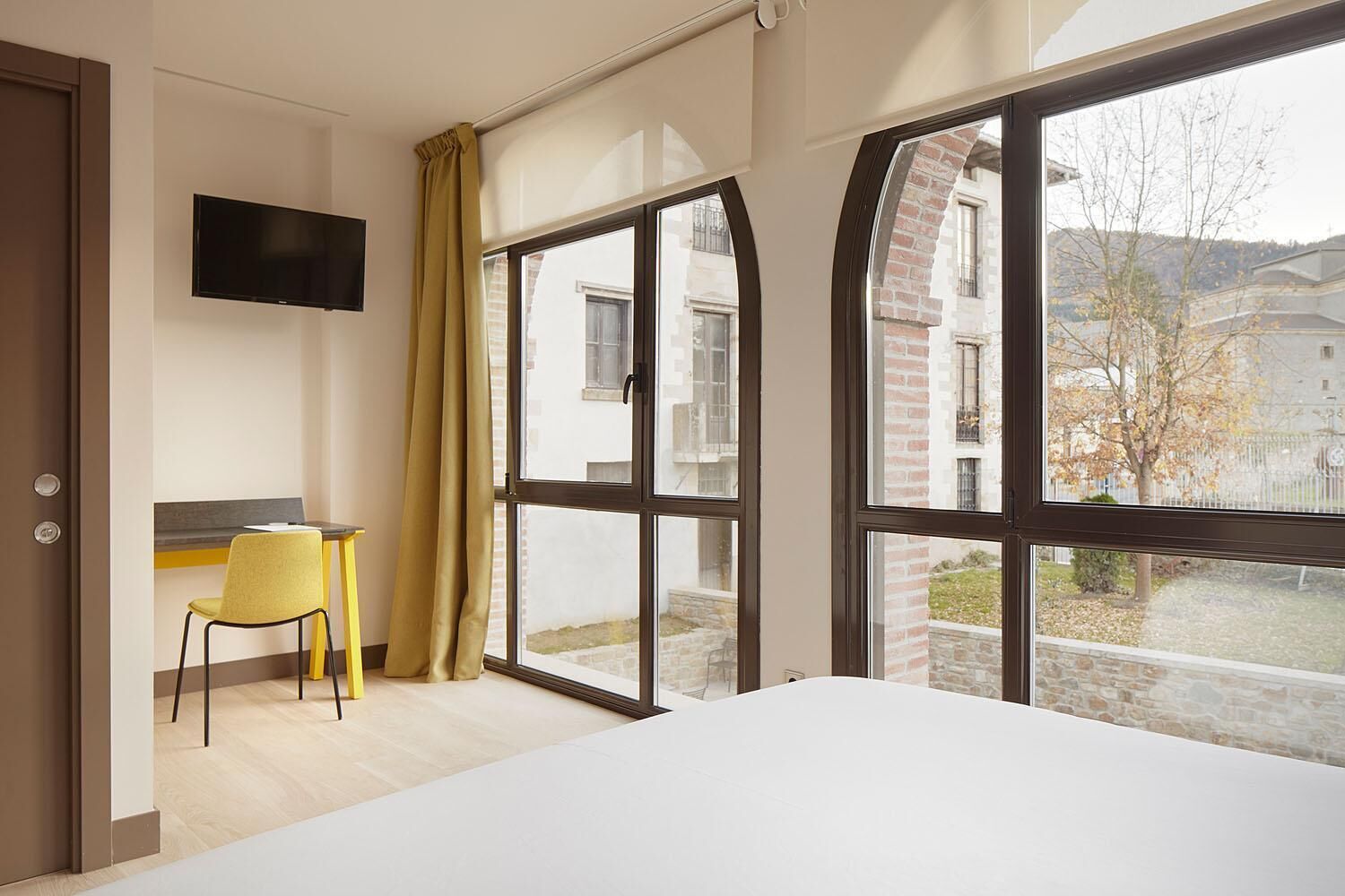 A hotel room with a bed, a desk with a chair, a TV mounted on the wall, and large windows with a view of buildings and trees.