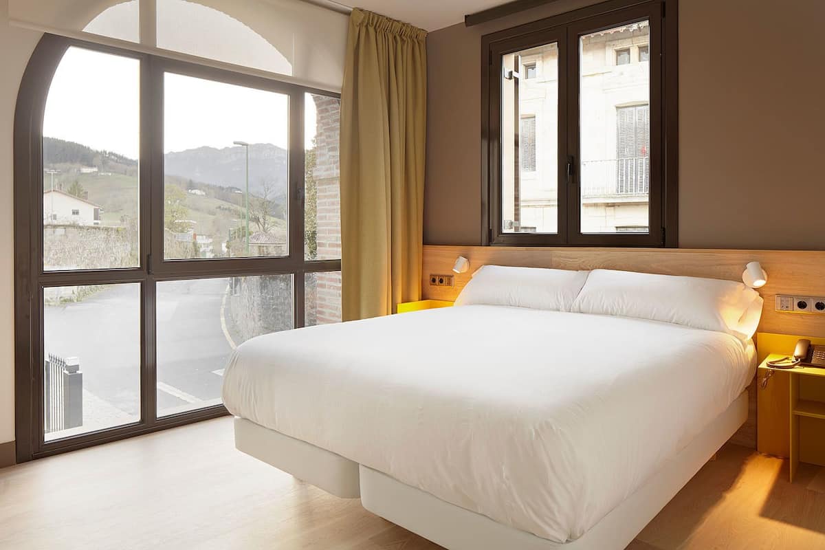 Ein Hotelzimmer mit einem großen Bett, zwei Nachttischen, einem Fenster mit Vorhängen und Blick nach draußen.