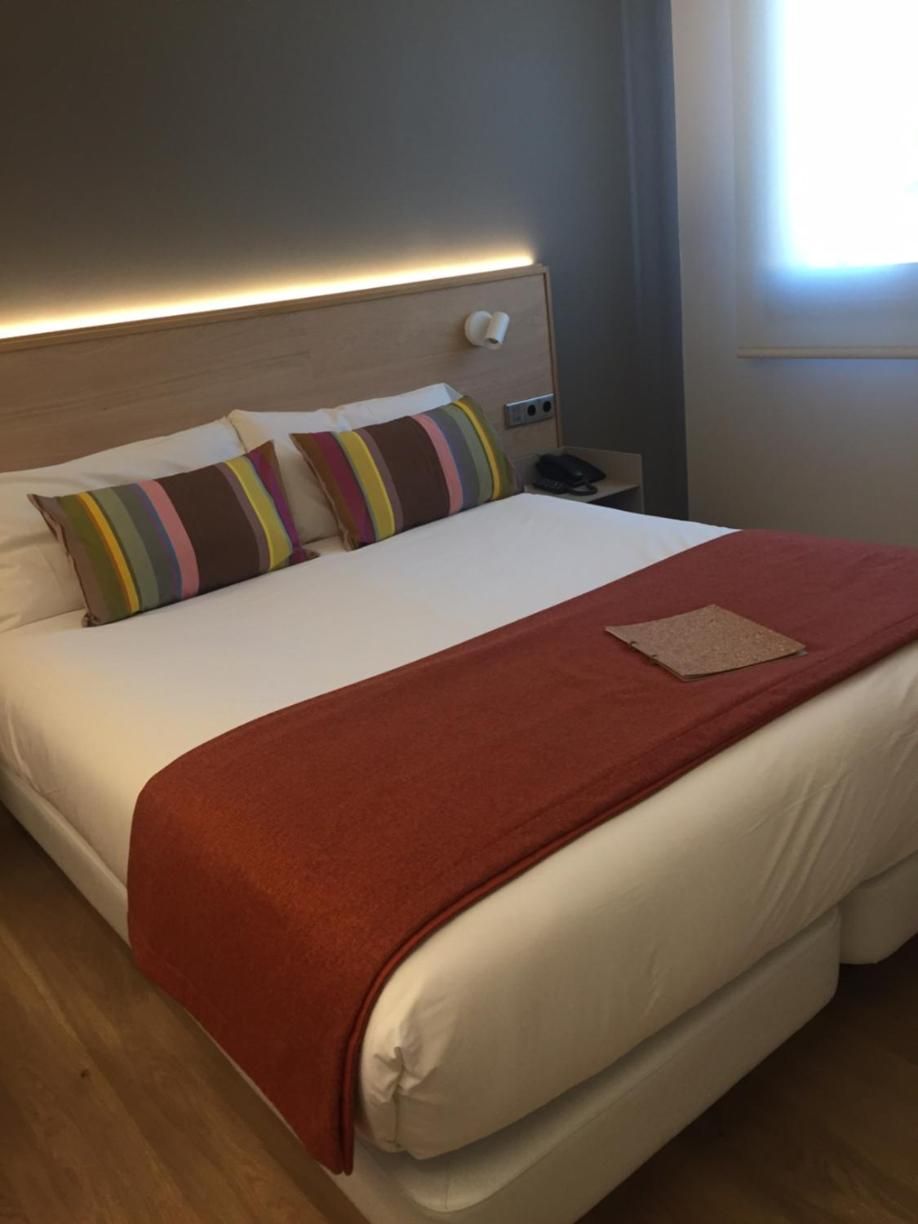 A hotel room with a bed, pillows, a nightstand, and a window.