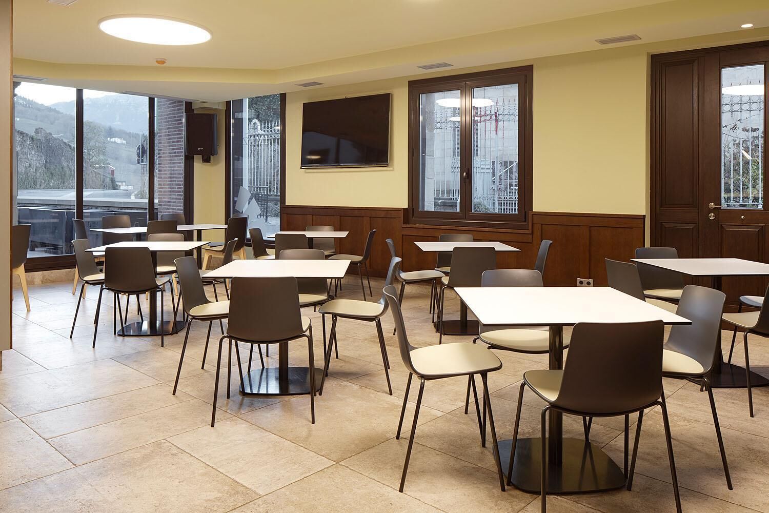 A dining area with multiple tables and chairs, large windows with a view of buildings, and a flat-screen TV mounted on the wall.