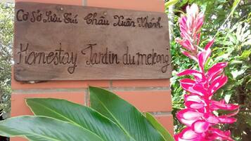 A wooden sign with text in a foreign language, a large green plant, and a pink flower.