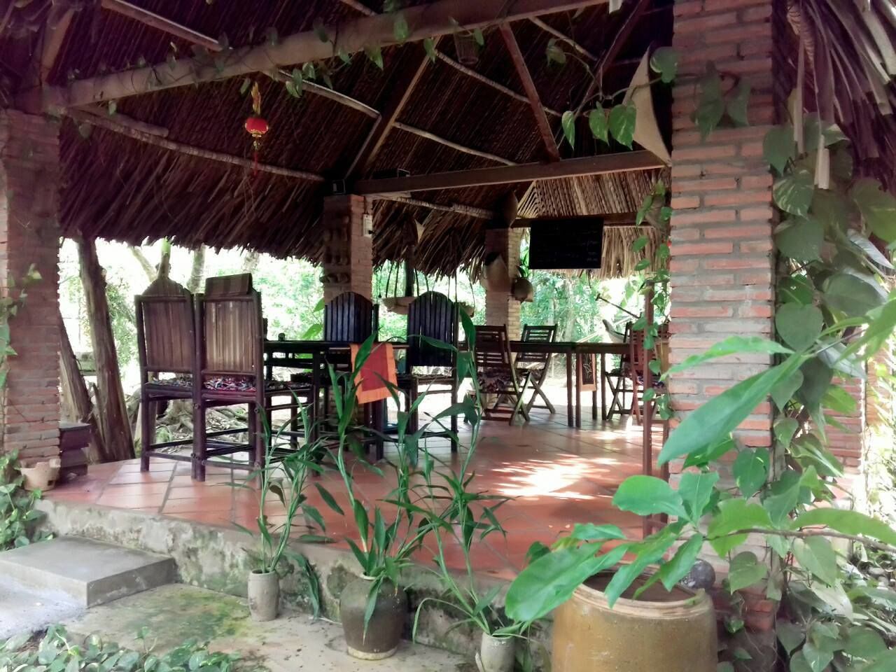A covered outdoor seating area with brick walls and a thatched roof, surrounded by potted plants.