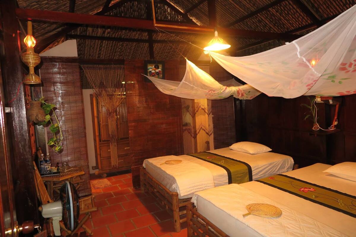 Una habitación tradicional con dos camas, piso de madera, techo de paja y mosquitero colgante.