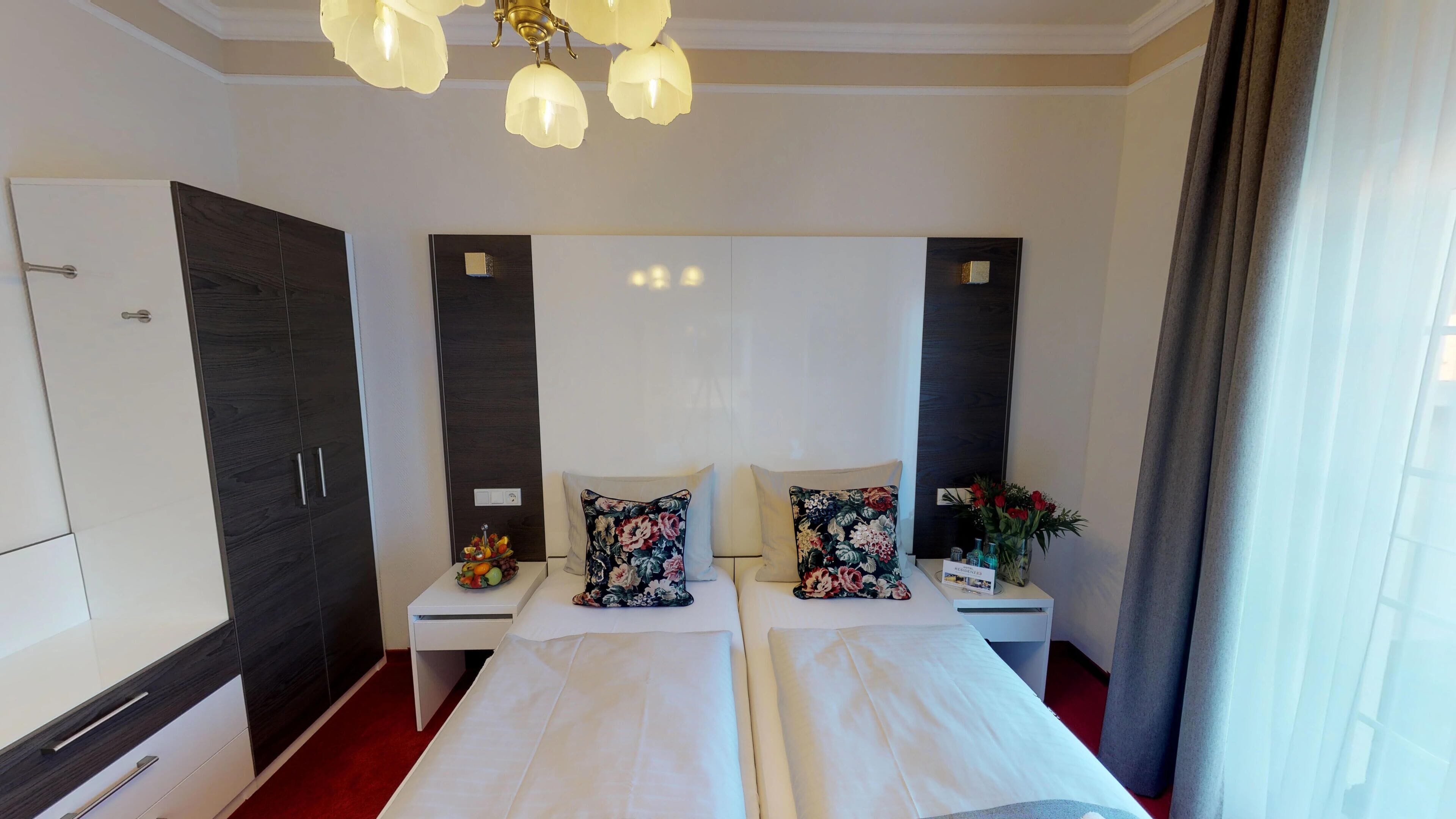 A hotel room with two beds, a wardrobe, a nightstand with a fruit bowl, and a decorative flower arrangement.