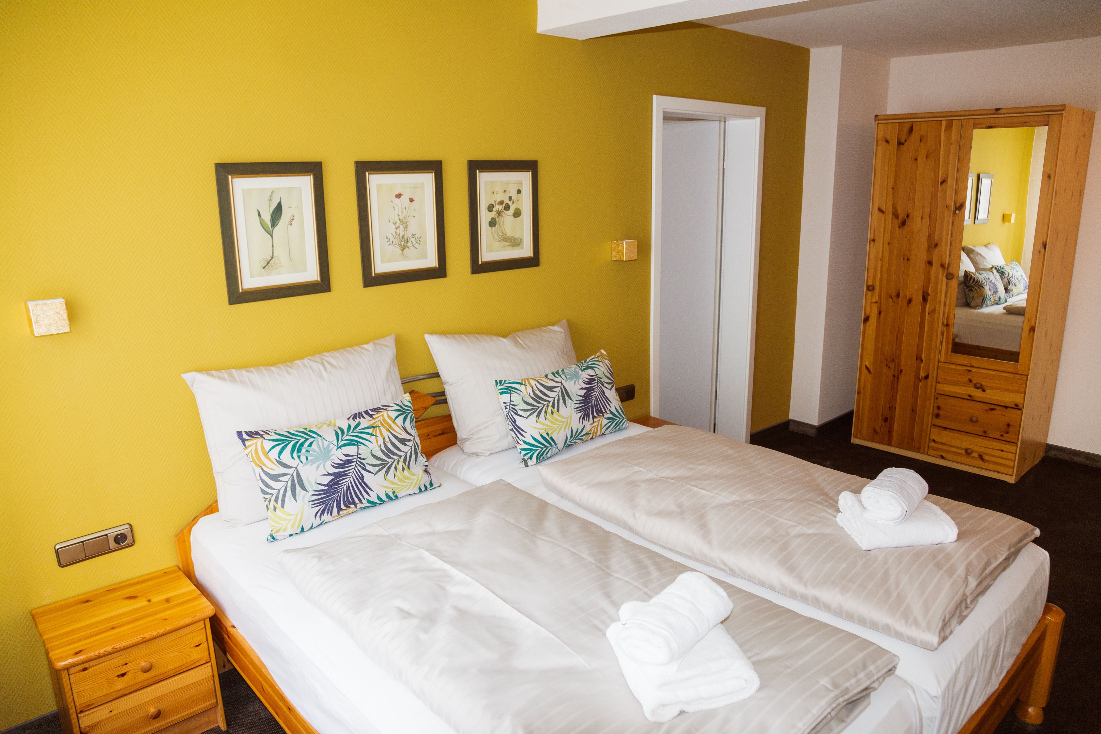 A double bed with white linens and tropical patterned pillows, a wooden nightstand, and a wooden wardrobe with a mirror.