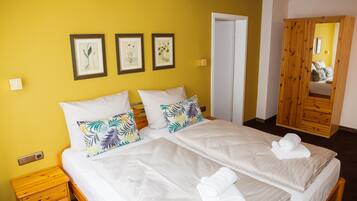 A double bed with white linens and tropical patterned pillows, a wooden nightstand, and a wooden wardrobe with a mirror.