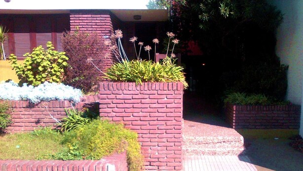 A brick planter box with plants and flowers, a brick wall, and a paved walkway. - Posada Galicia (Villa Maria)