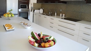 A modern kitchen with white cabinets, a built-in oven, microwave, and a large island with bowls of fruit. - Elina Luxury Residence Villa (Chania)