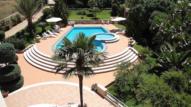 A pool surrounded by a circular deck with lounge chairs and umbrellas, set in a lush garden with palm trees and a view of distant mountains. - Elina Luxury Residence Villa (Chania)