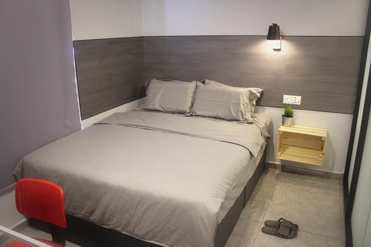 A neatly made bed with a grey headboard, a small bedside table with a plant, and a red chair.