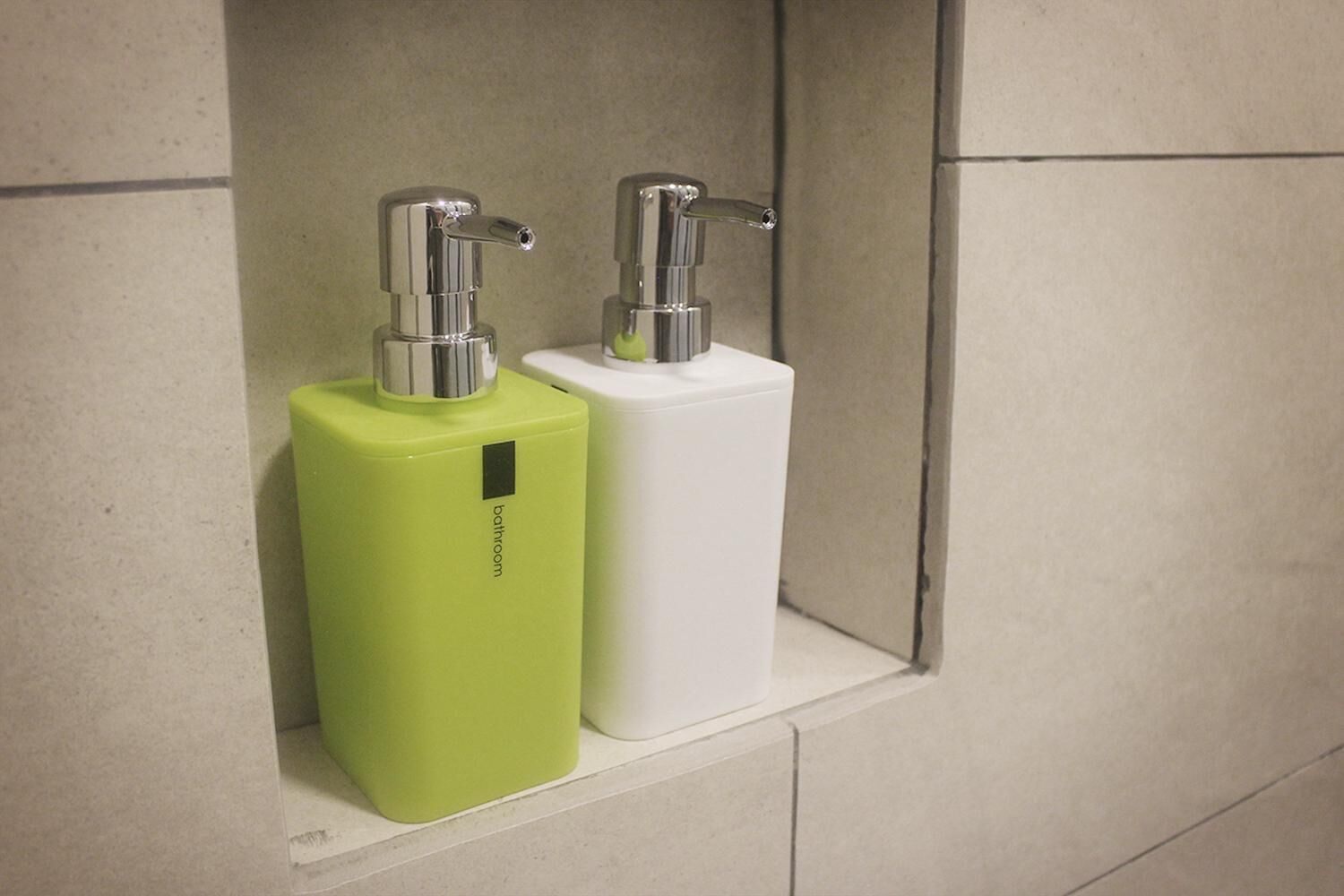 Dos dispensadores de baño, uno verde y otro blanco, sujetos a una pared con azulejos.