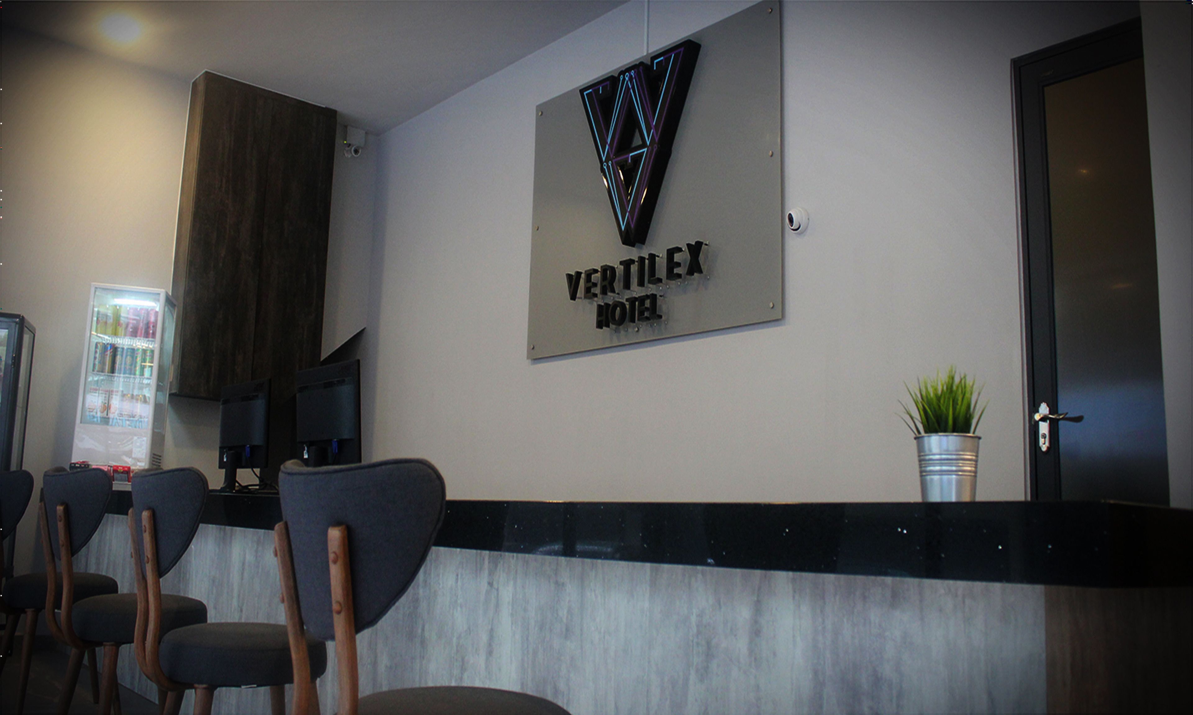 A reception area with a sign reading "Vertilex Hotel," a refrigerator, a plant, and several chairs.