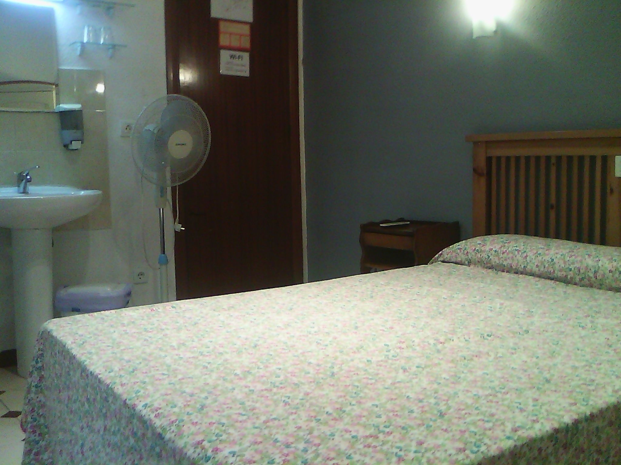 Habitació d'hotel amb llit, una taula de nit, un panell de capçalera de fusta, un ventilador fixat a la paret, un rentanet i un mirall.